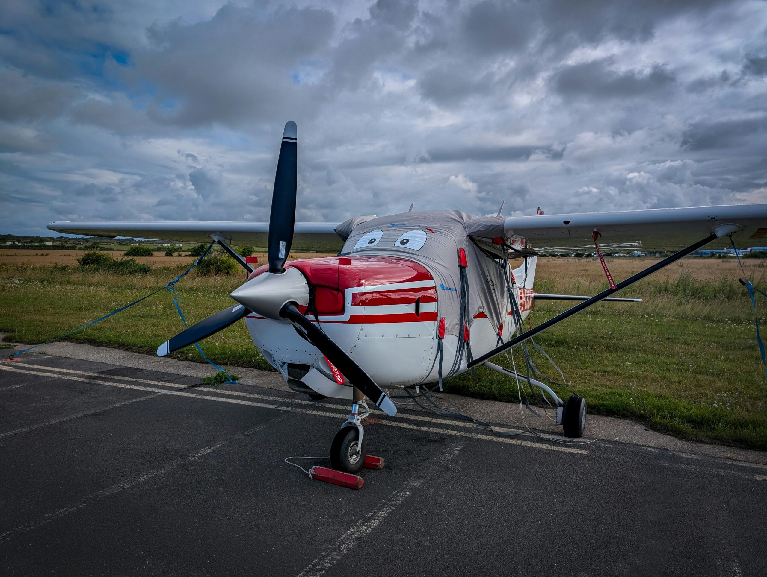 A small red and white airplane parked on an airstrip on a cloudy day, with a protective cover on the nose.